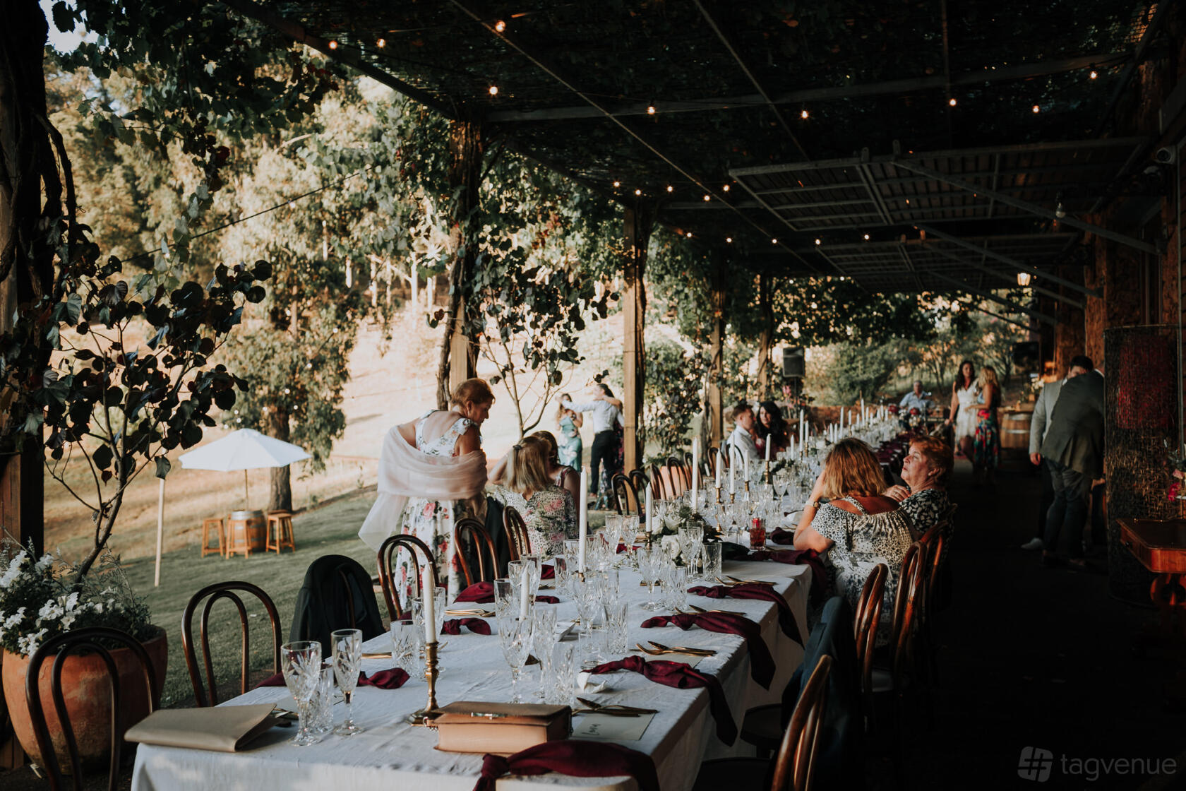 An outdoor terrace with long banquet tables, burgundy napkins, and overhead string lights at Stonebarn Weddings.