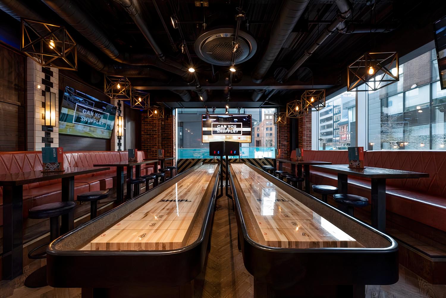 A bar with shuffleboard tables, red leather banquette seating, and geometric pendant lights at Box Deansgate.