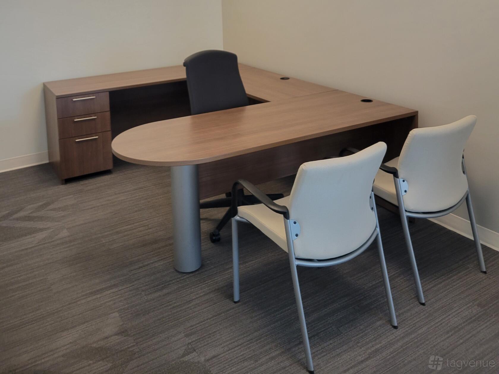 An office space with a large L-shaped wooden desk, two guest chairs, and neutral carpeting at Coalition Space Denver.