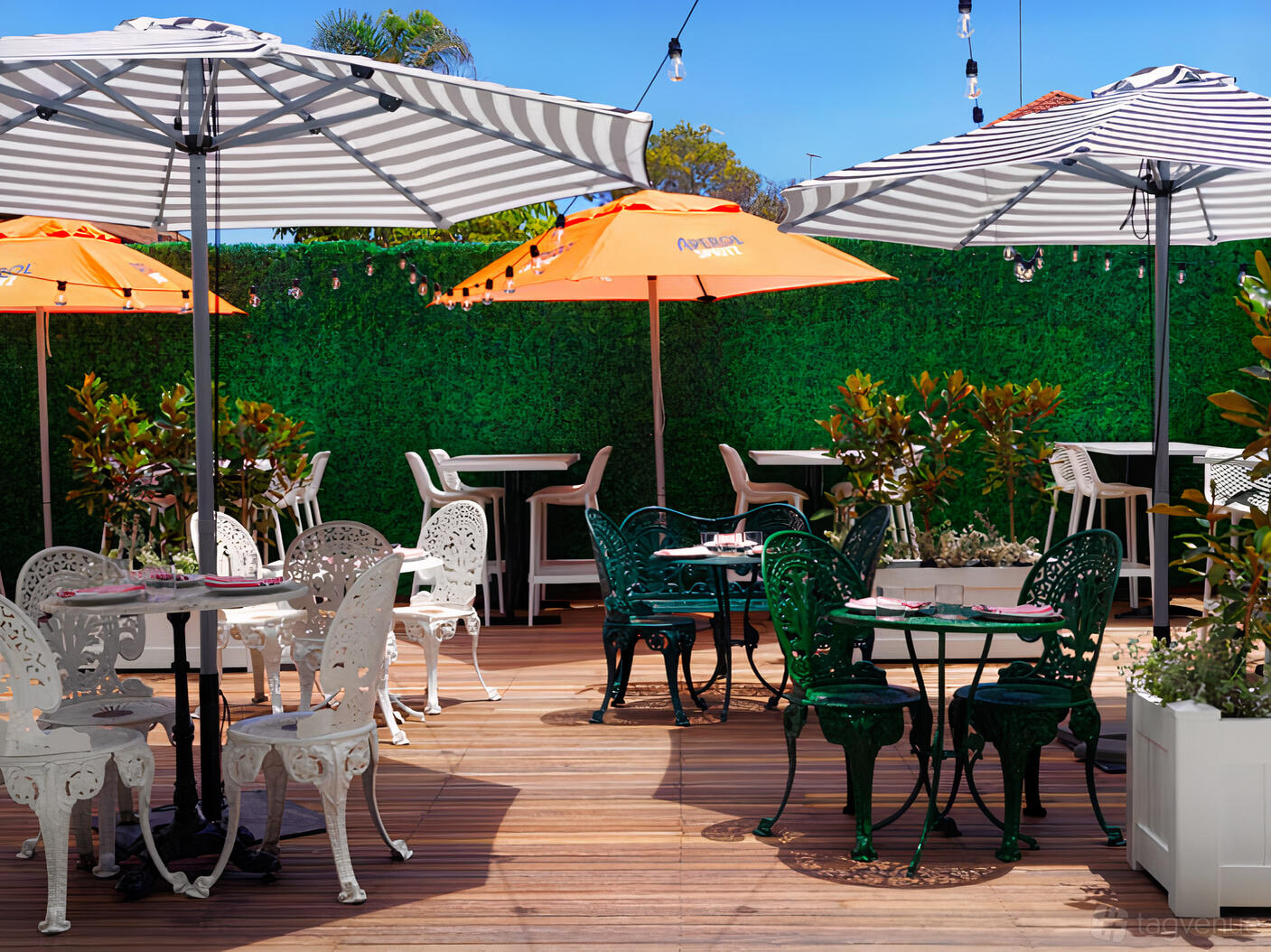 A restaurant terrace with striped and orange umbrellas, metal bistro tables, and greenery along the fence at barmilano.