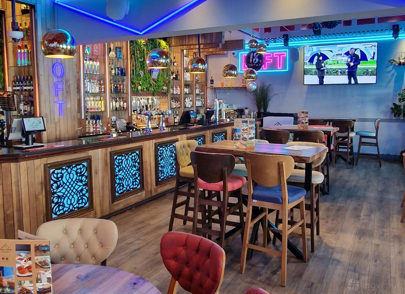 An event space in a bar with a long wooden counter, backlit shelves, colorful chairs, and pendant lighting at THE LOFT WOOLTON.