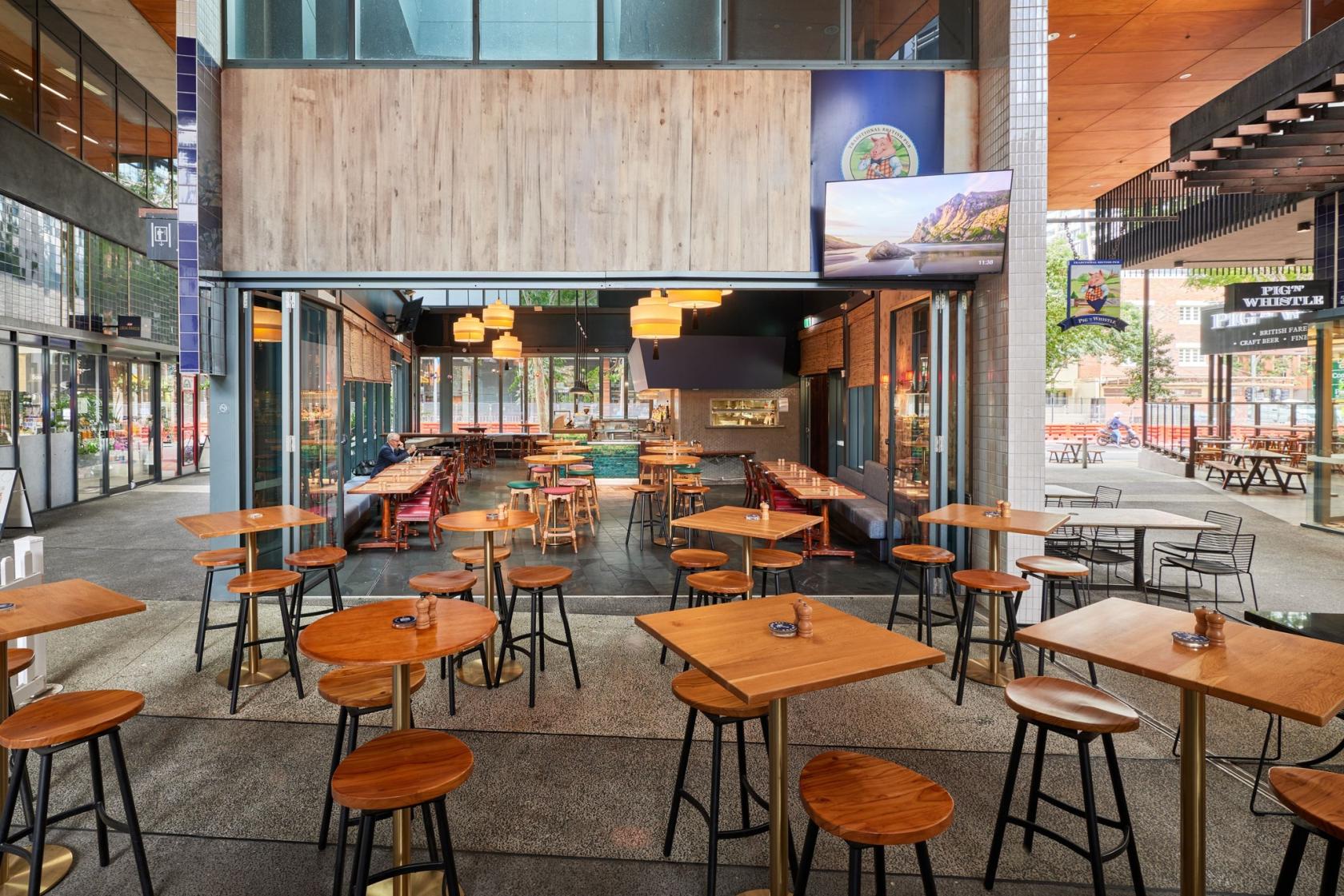An event space in a restaurant with high-top wooden tables, stools, and large glass windows at Pig ‘N’ Whistle - West End.