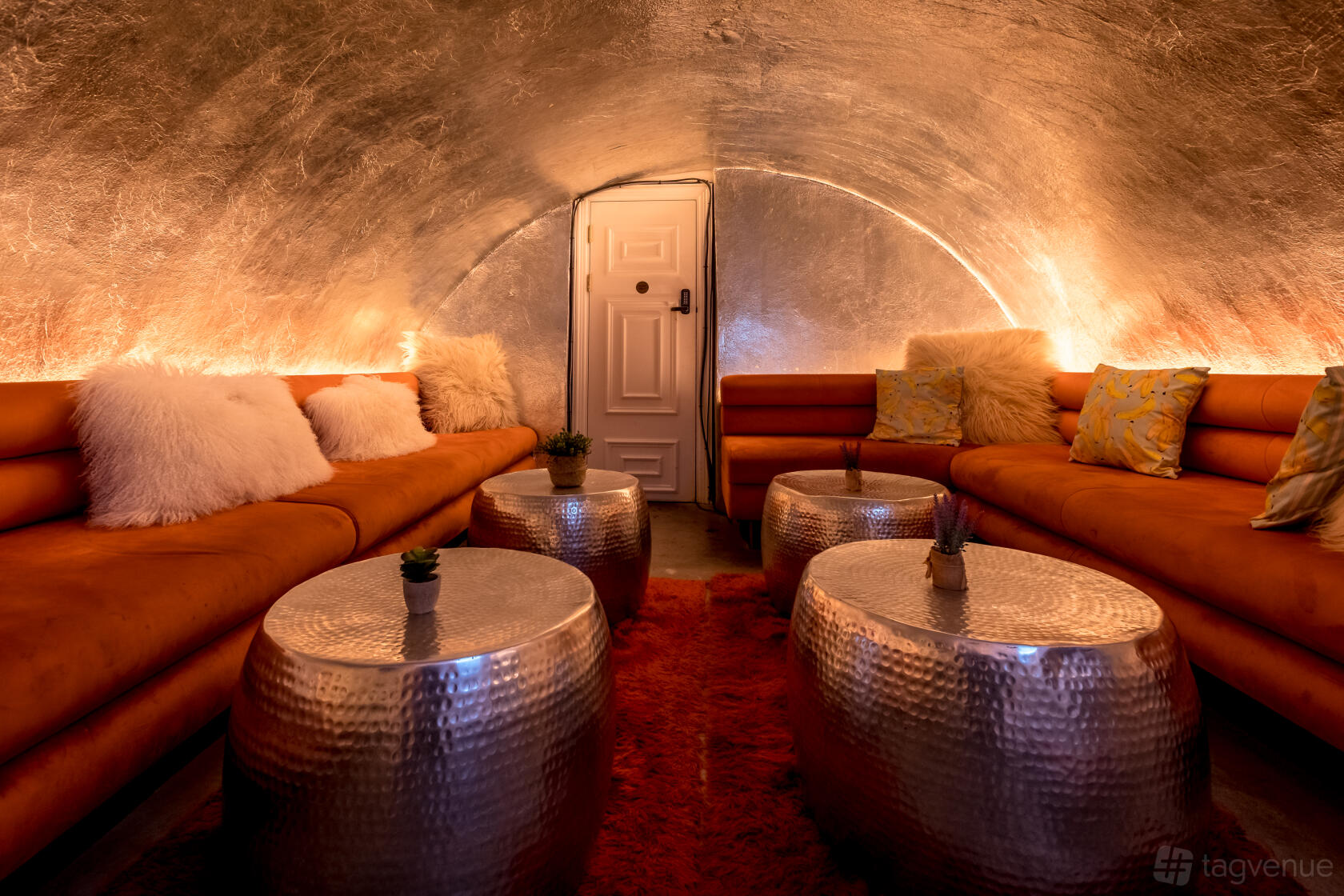 A cocktail bar lounge with curved metallic walls, plush sofas, and round hammered metal tables at The Little Scarlet Door.