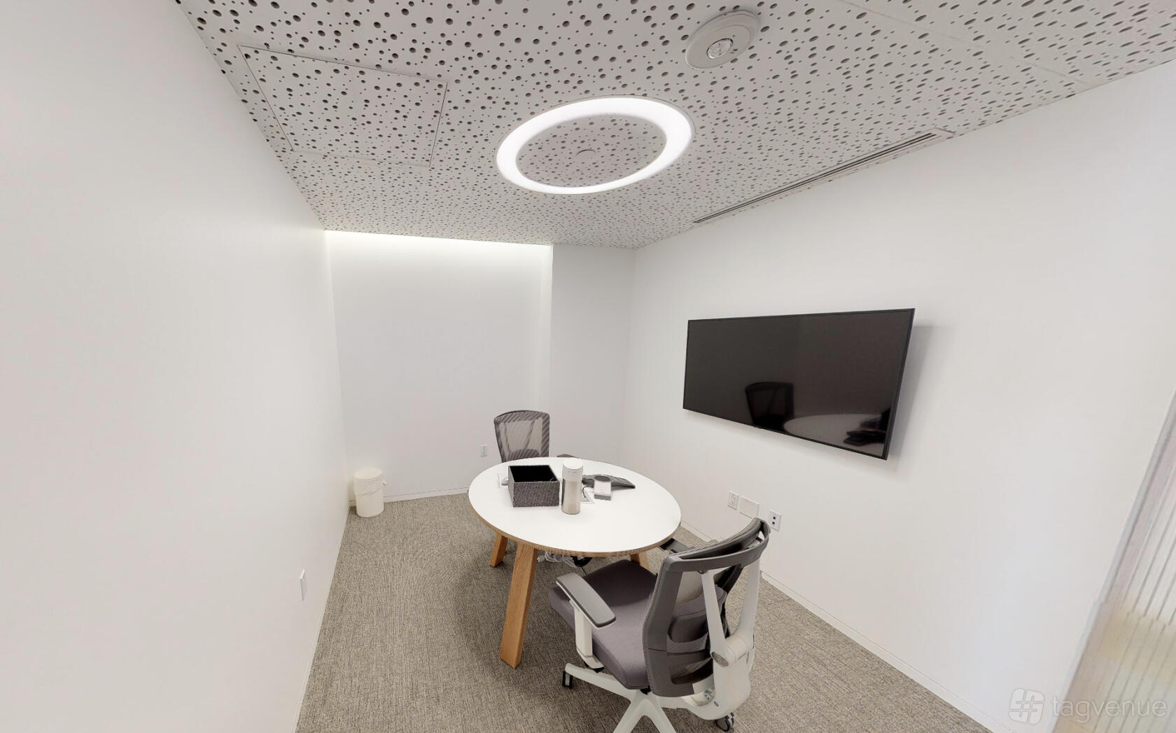 A meeting room with a round table, two mesh chairs, wall-mounted screen, and white patterned ceiling at Ethos Society Koreatown.