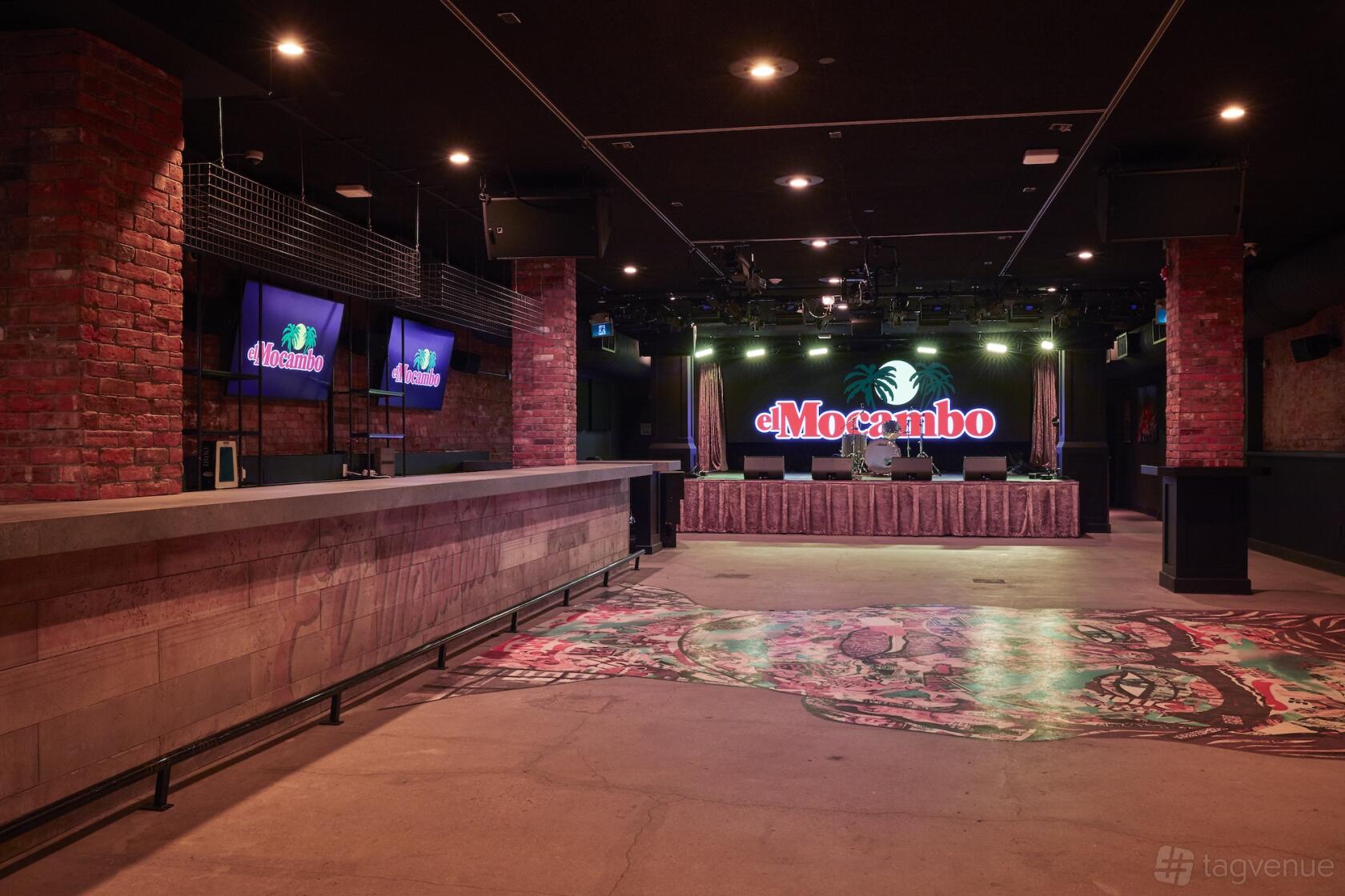 An event venue with exposed brick columns, a long bar counter, and a stage with LED signage at El Mocambo.