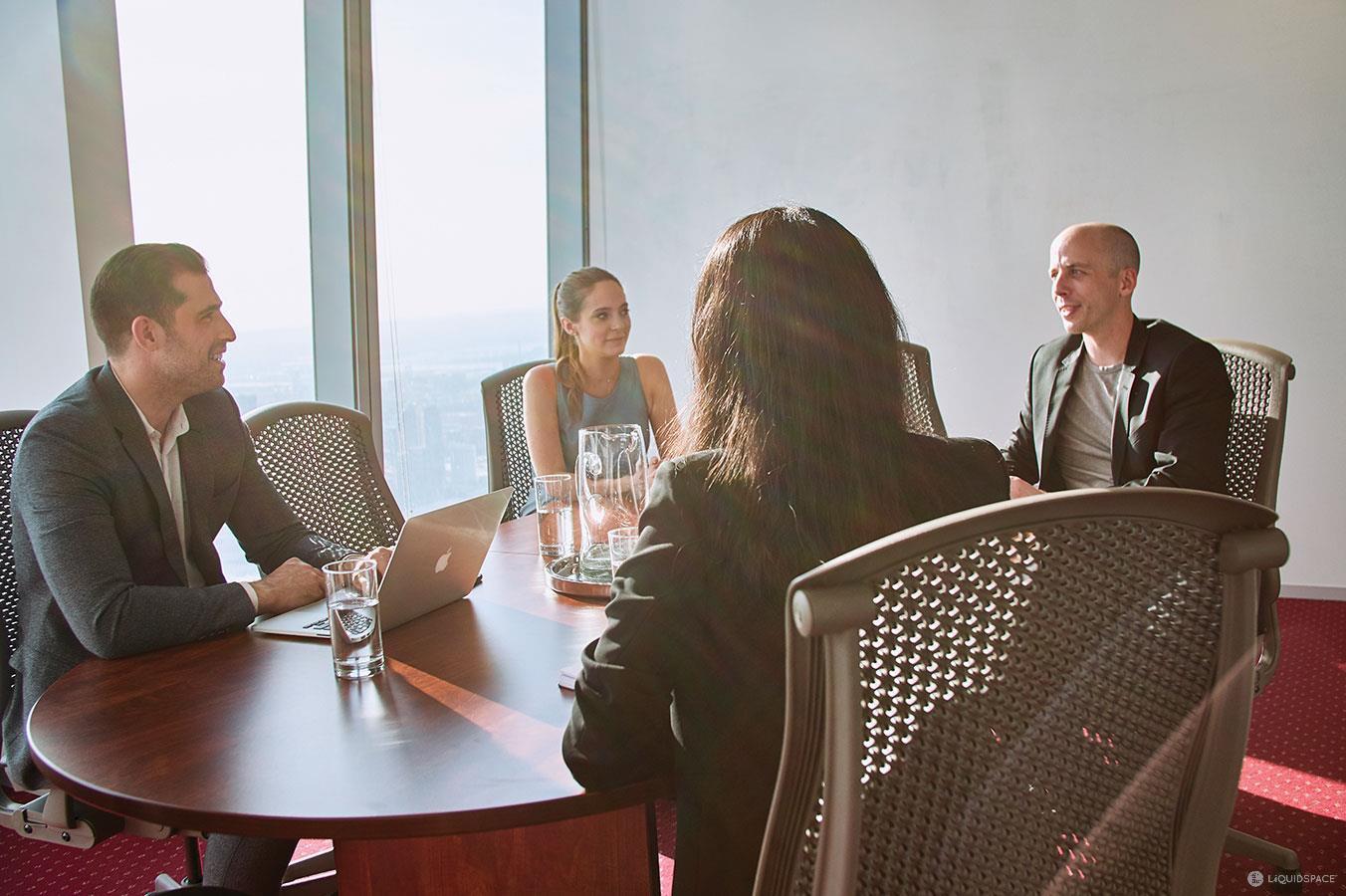 A meeting room with a round wooden table, mesh chairs, and floor-to-ceiling windows at Servcorp - The Loop, Chicago.