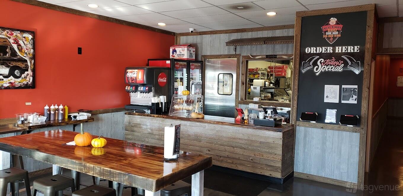 A restaurant interior with a wooden counter, soda fountain, menu board, and communal table at MeatWagon BBQ.