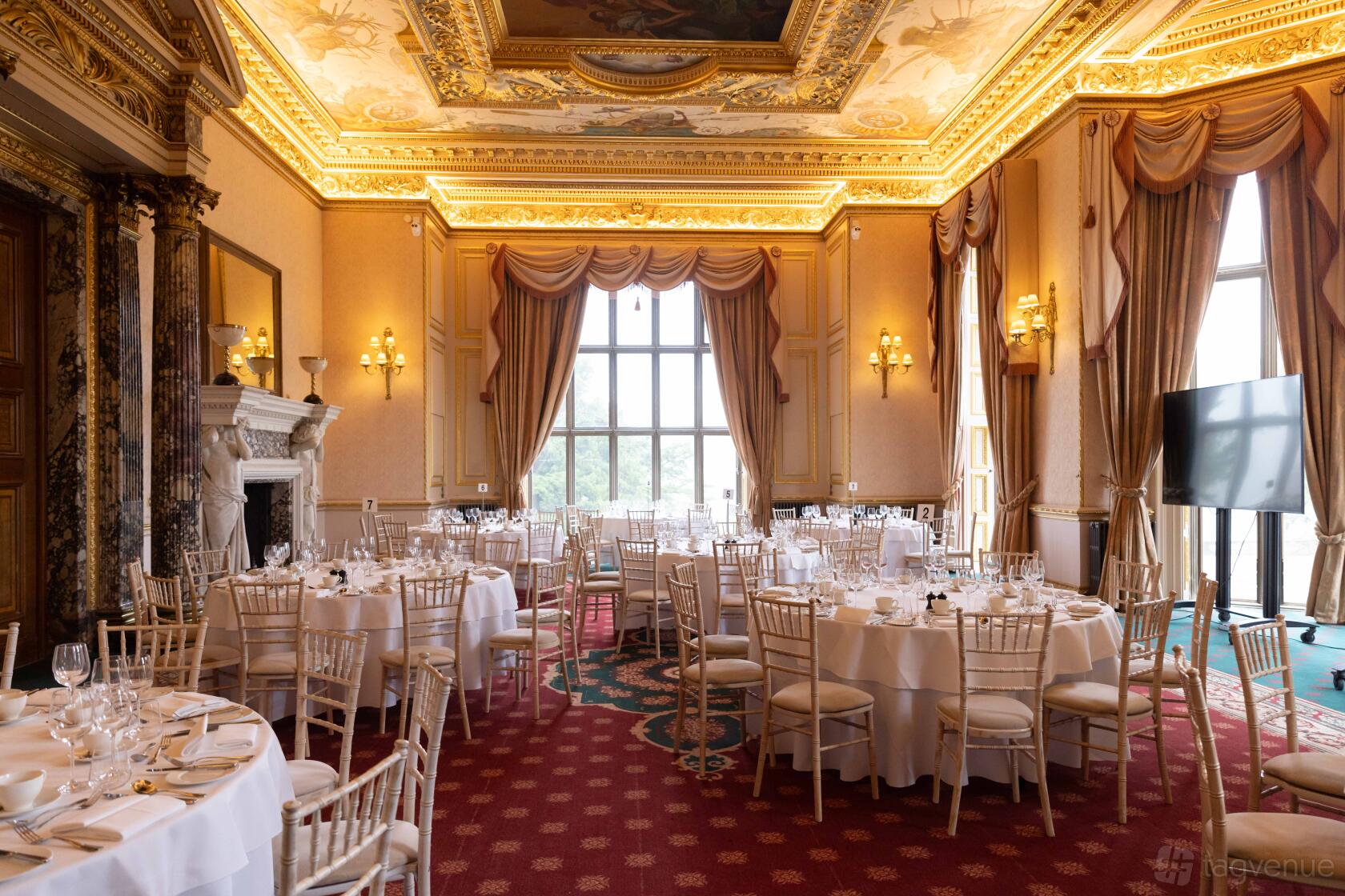 An ornate event space with gilded ceiling, tall windows, and round tables with white linens at Ashridge House.