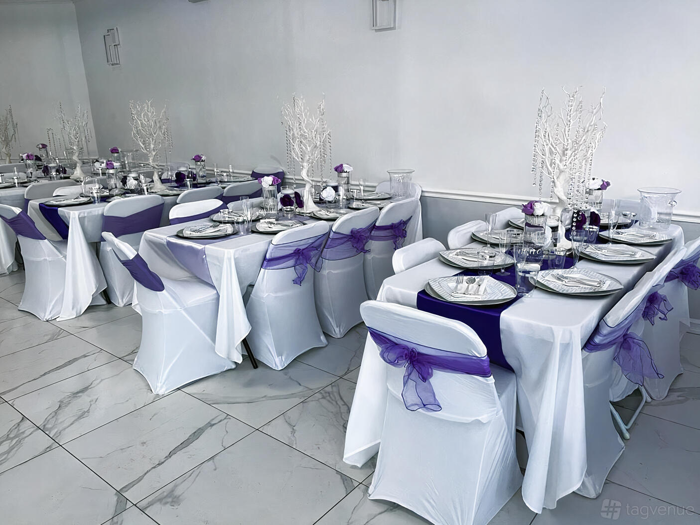 An event venue with rectangular tables dressed in white linens, purple accents, and white branch centerpieces at Strawberry Events.