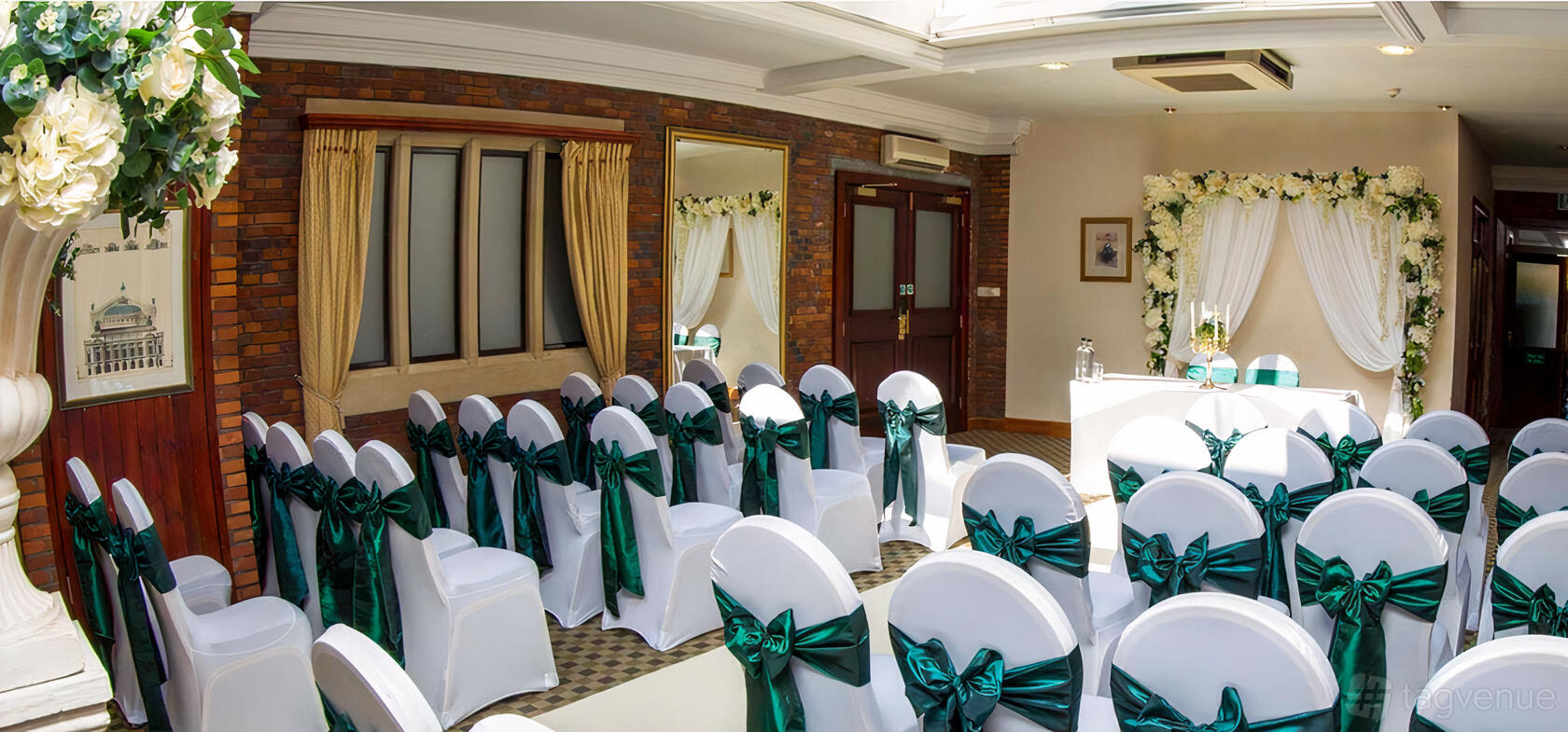 An event suite with rows of white chairs with green sashes, brick walls, gold curtains, and floral arches at Dunston Hall.