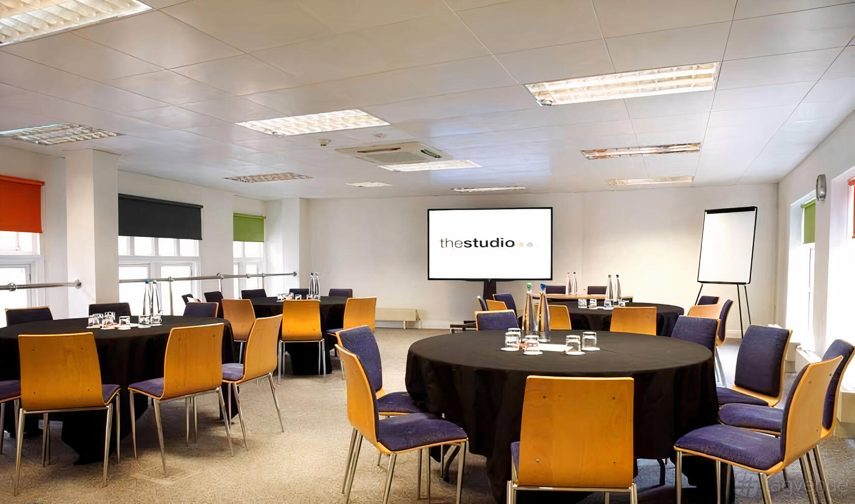 A meeting centre with round tables, wooden chairs, a projector screen, and flipchart at The Studio Birmingham.