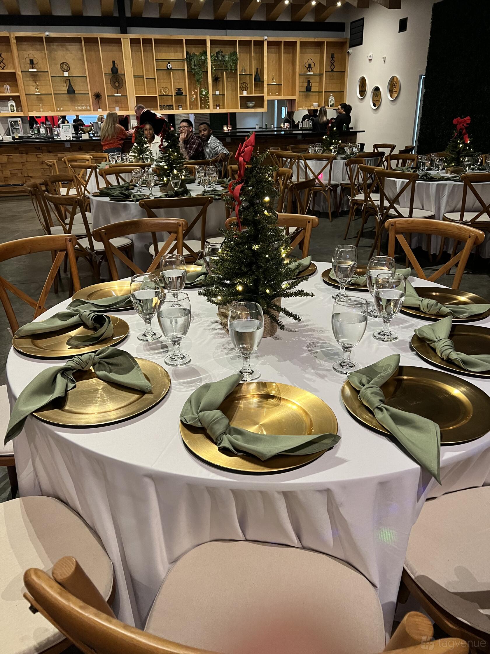 An event venue with round tables set with gold chargers, white linens, green napkins, and a small decorated tree centerpiece at La Galleria Venue.