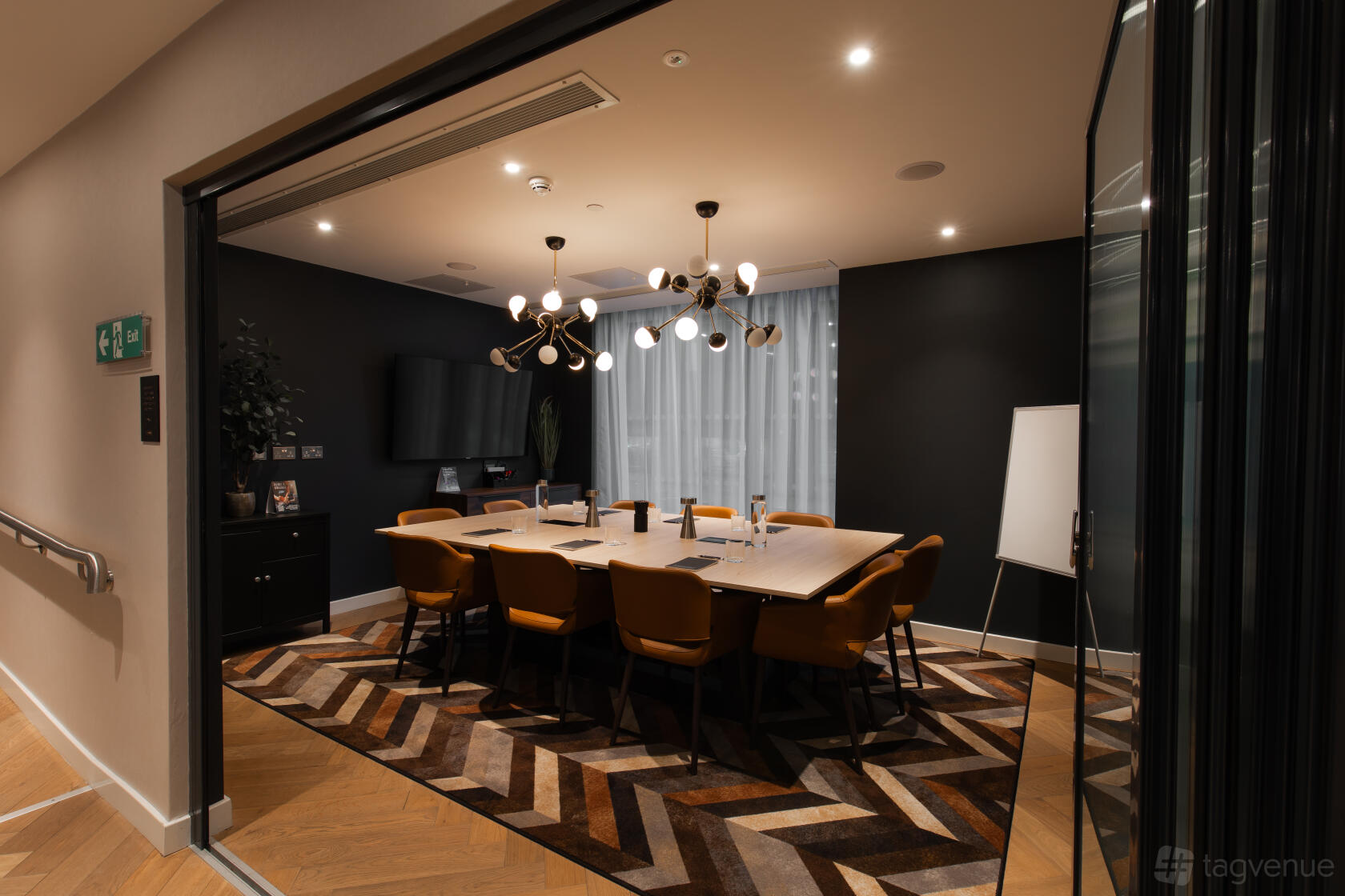 A hotel meeting room with a long table, orange chairs, modern chandelier, and geometric rug at CitySuites.