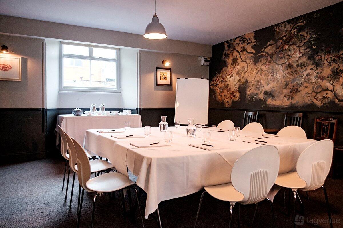 A dining room with white-clothed tables arranged in a square, white chairs, and a mural wall at The Bull Hotel Dorset.