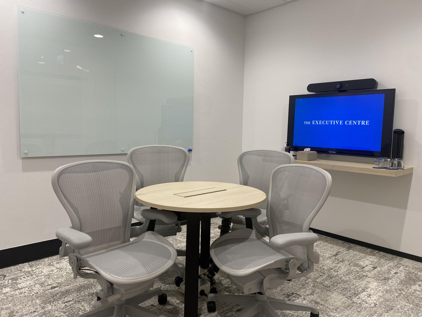 A meeting room with a round table, four mesh office chairs, a wall-mounted screen, and a glass whiteboard at Angel Place.