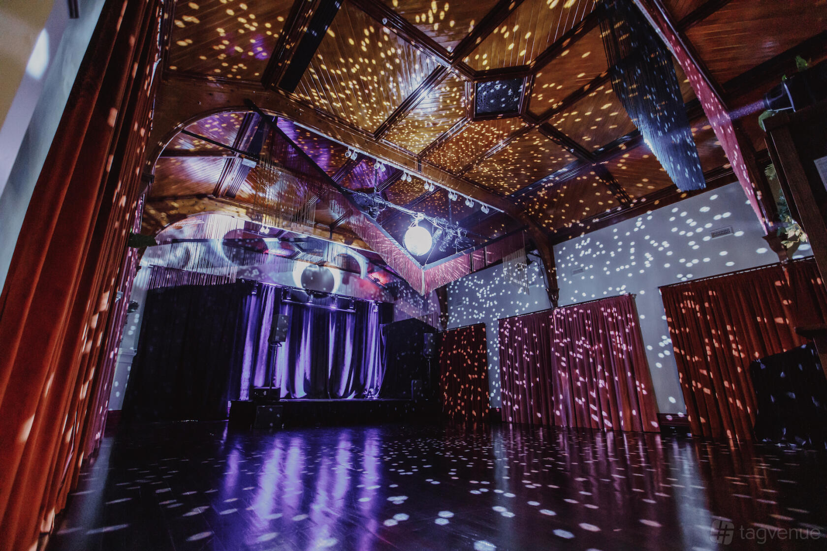 A hall with wood-paneled vaulted ceilings, stage, red curtains, and disco lighting at Fringe Common Rooms.
