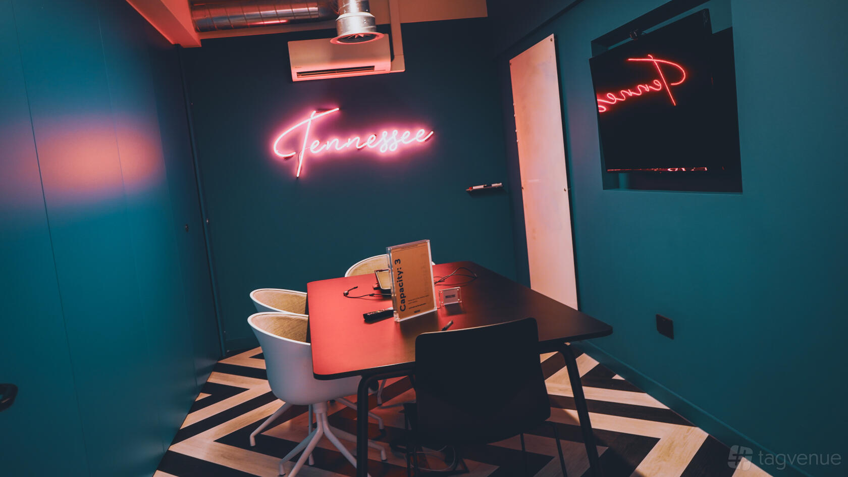 A meeting room with teal walls, a chevron-patterned floor, neon Tennessee sign, and white chairs at Huckletree Ancoats.