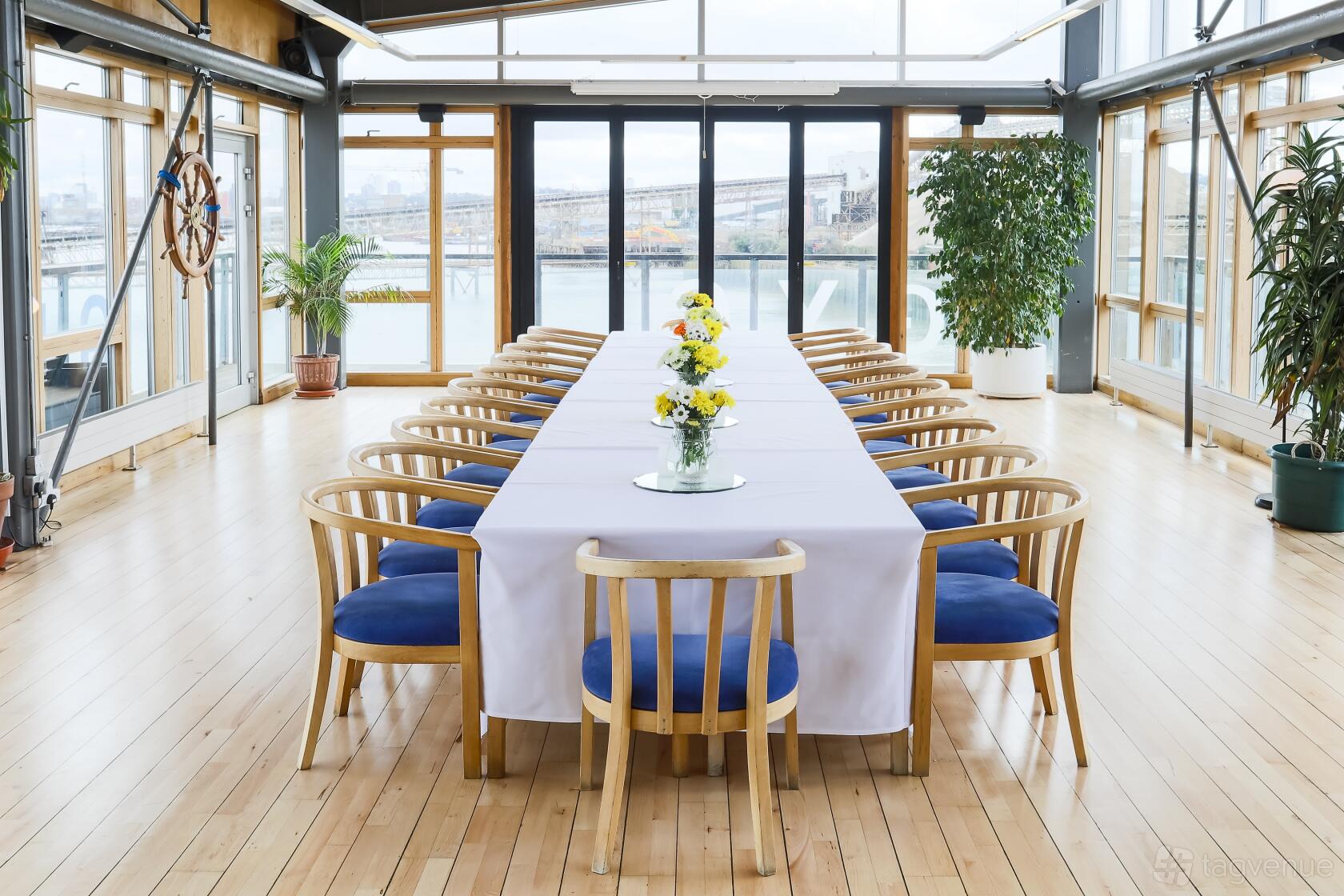 An event venue with floor-to-ceiling windows, potted plants, and a long table with white linens at Greenwich Yacht Club.