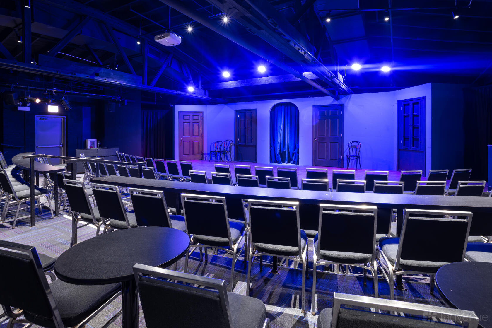 An auditorium with black chairs, long tables, a small stage, and blue theatrical lighting at iO Theater.