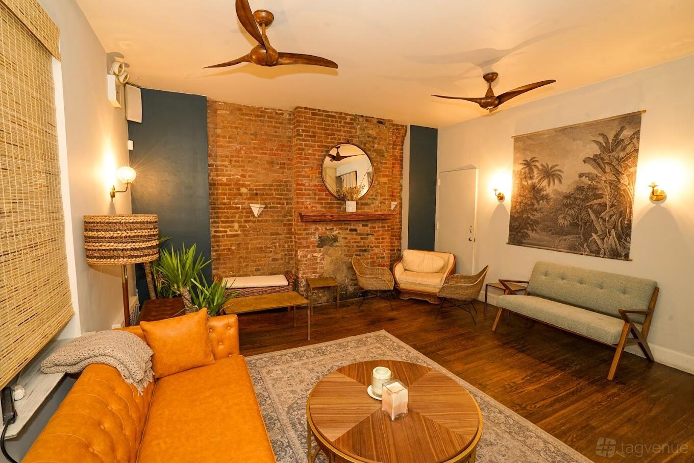 A private dining room with exposed brick wall, round wooden coffee table, tan leather sofa, and mid-century chairs at Ama Raw Bar East Village.