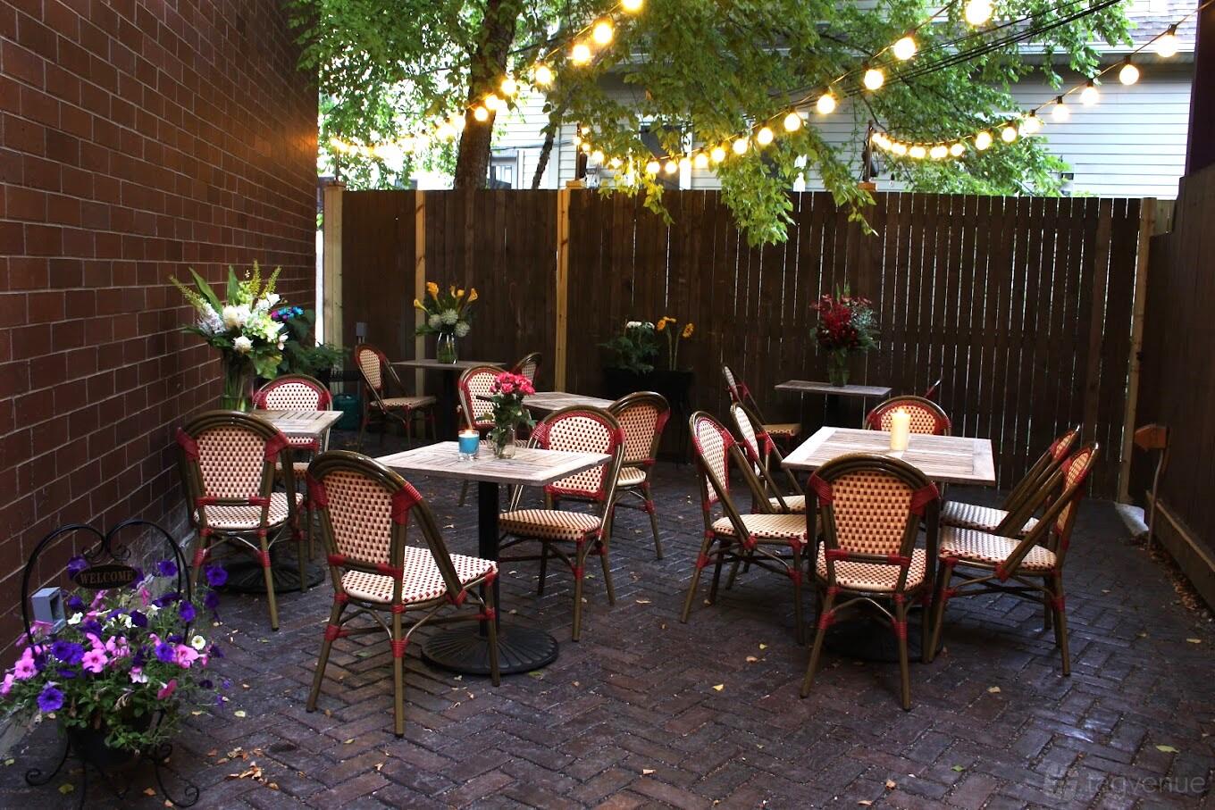 An outdoor restaurant patio with bistro chairs, round tables, string lights, and surrounding greenery at Cobblestone.