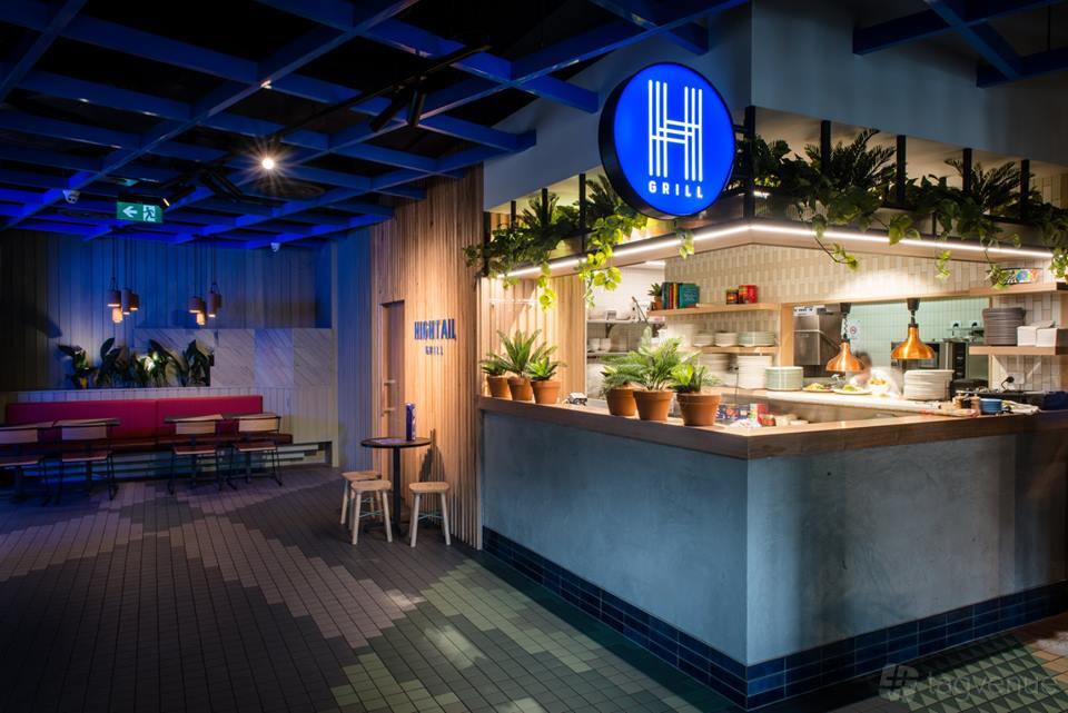 A bar with an open kitchen, potted plants, and booth seating under blue ceiling beams at Hightail.