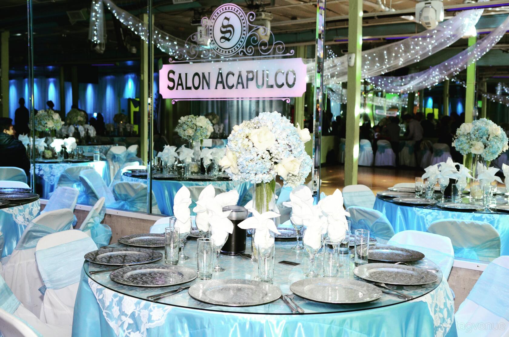 A banquet hall with mirrored walls, round tables dressed in blue linens, and floral centerpieces at Salon Acapulco Banquet Hall.