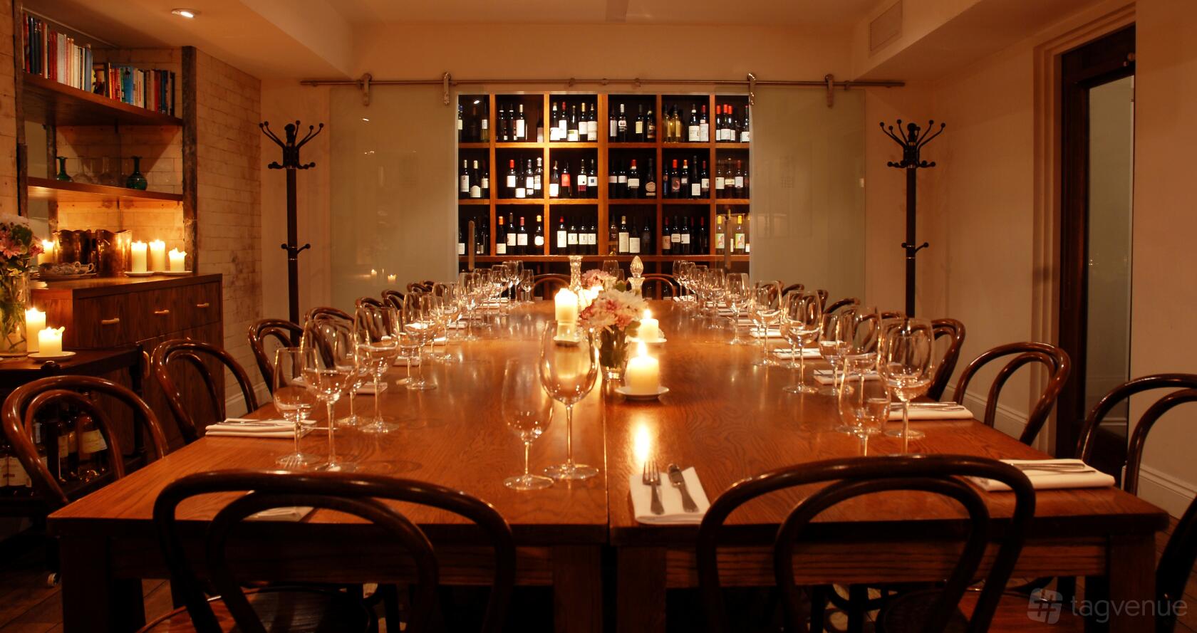 A dining room with a long wooden table set with glassware and candles, wine bottle shelves at Vinoteca Farringdon.