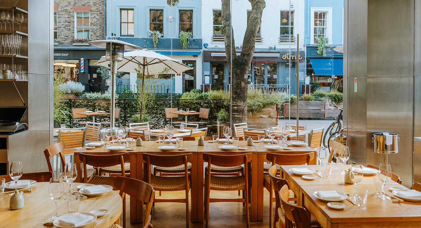 A restaurant with wooden tables set for group dining and floor-to-ceiling windows overlooking the street at Roka Charlotte Street.