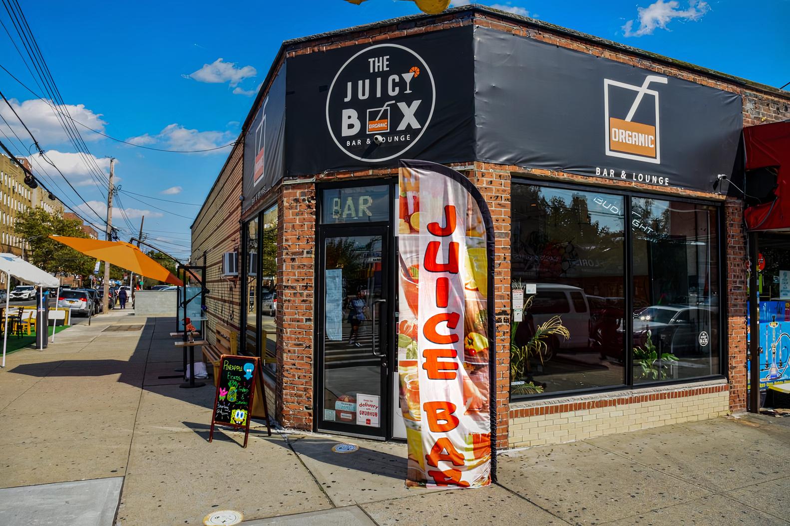 A corner bar with brick exterior, large black awning, and a colorful juice bar flag at The Juicy Box Bar.