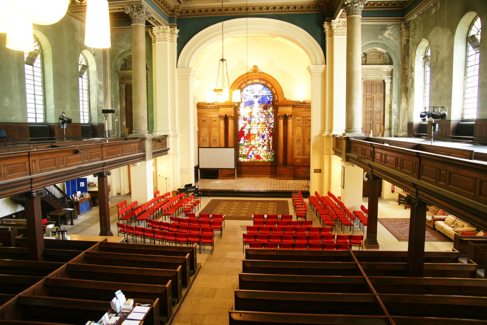 An event space in a church with red chairs, high ceilings, balcony seating, and a large stained glass window at St Anne's Limehouse.