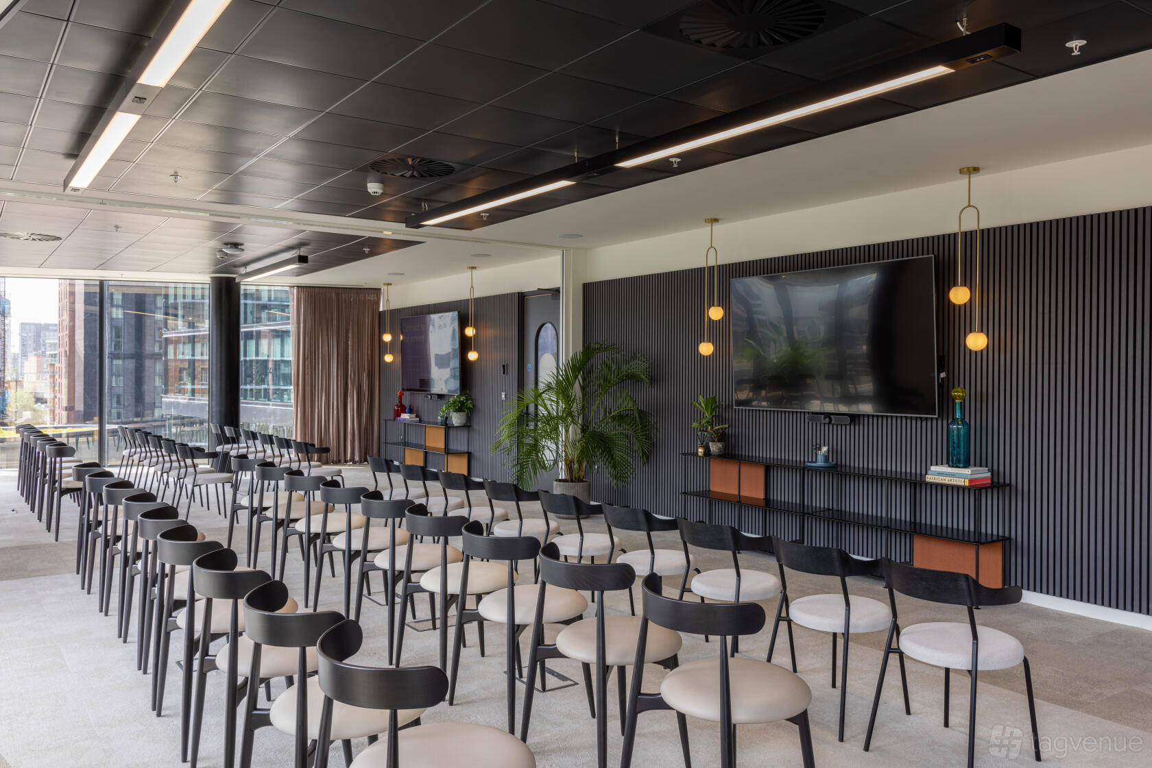 An event space with rows of black chairs, wall-mounted screens, and large windows at x+why - 100 Embankment.