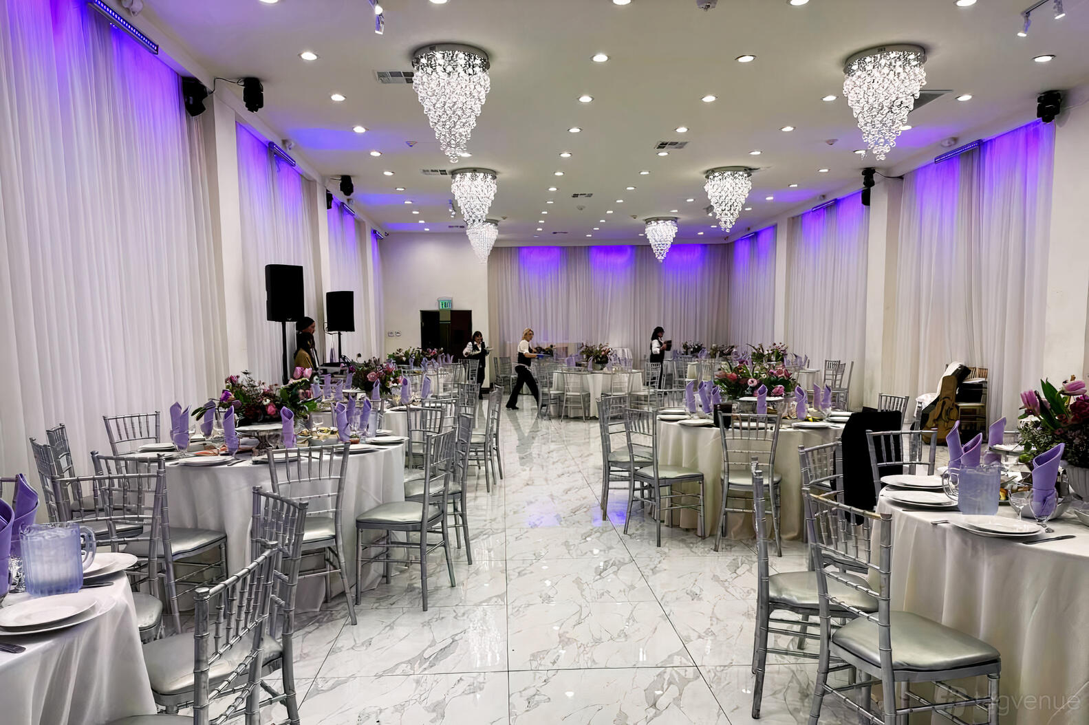 A hall with round tables set with floral centerpieces, silver chairs, marble floors, and crystal chandeliers at Vivaldi Venues.
