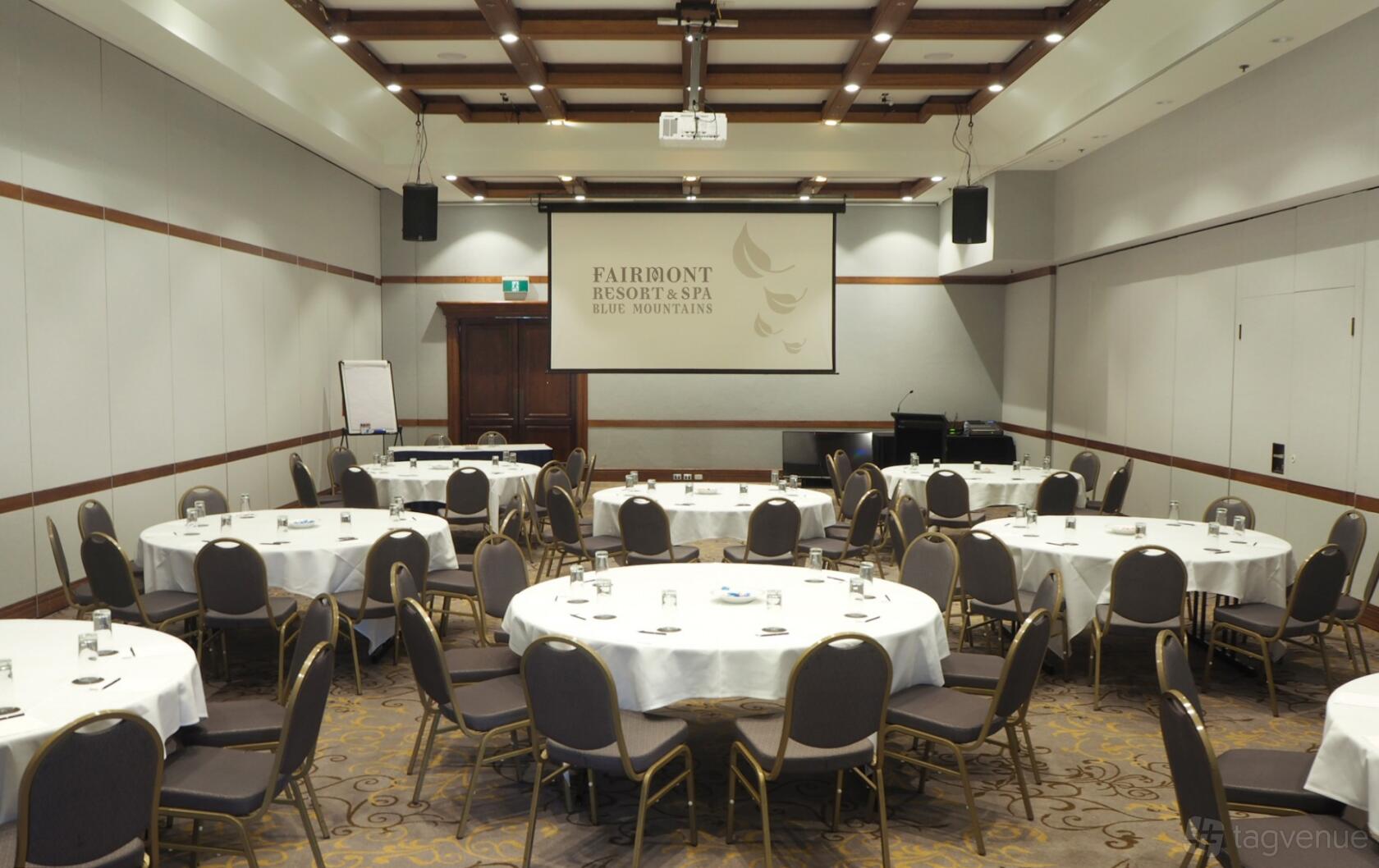 An event space with round tables draped in white linens, banquet chairs, and a ceiling-mounted projector at Fairmont Resort Blue Mountains.