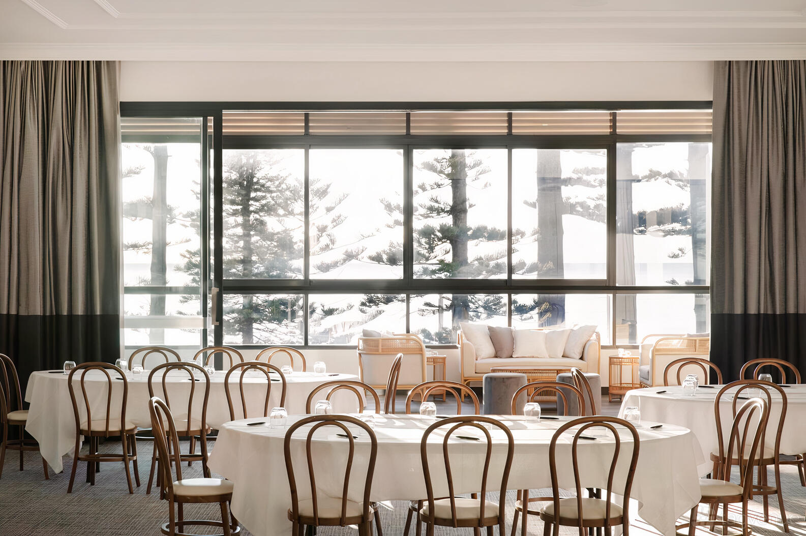 An event space with round tables draped in white linens, bentwood chairs, and large windows overlooking pine trees at Manly Pacific.