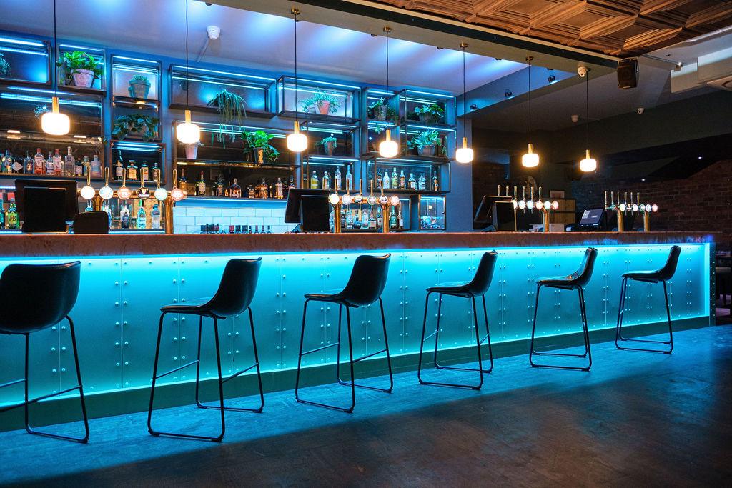 An event space in a karaoke bar with a long illuminated bar, high stools, and pendant lighting at Star by Liverpool Street.