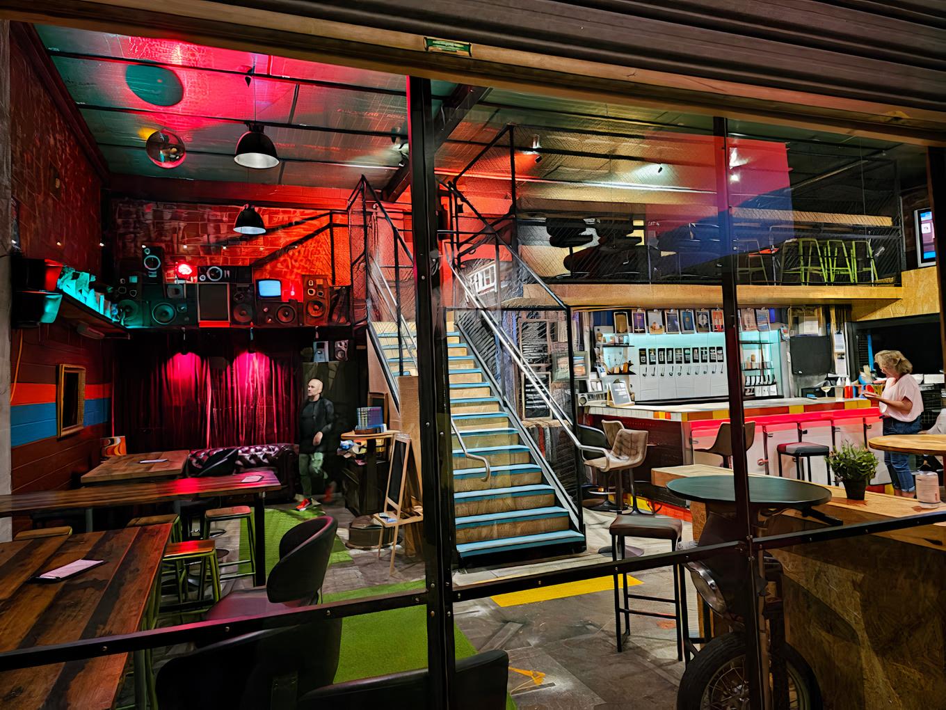 A taproom bar with colorful lighting, industrial decor, long wooden tables, and a central staircase at Liberty Brewing Co Taproom.