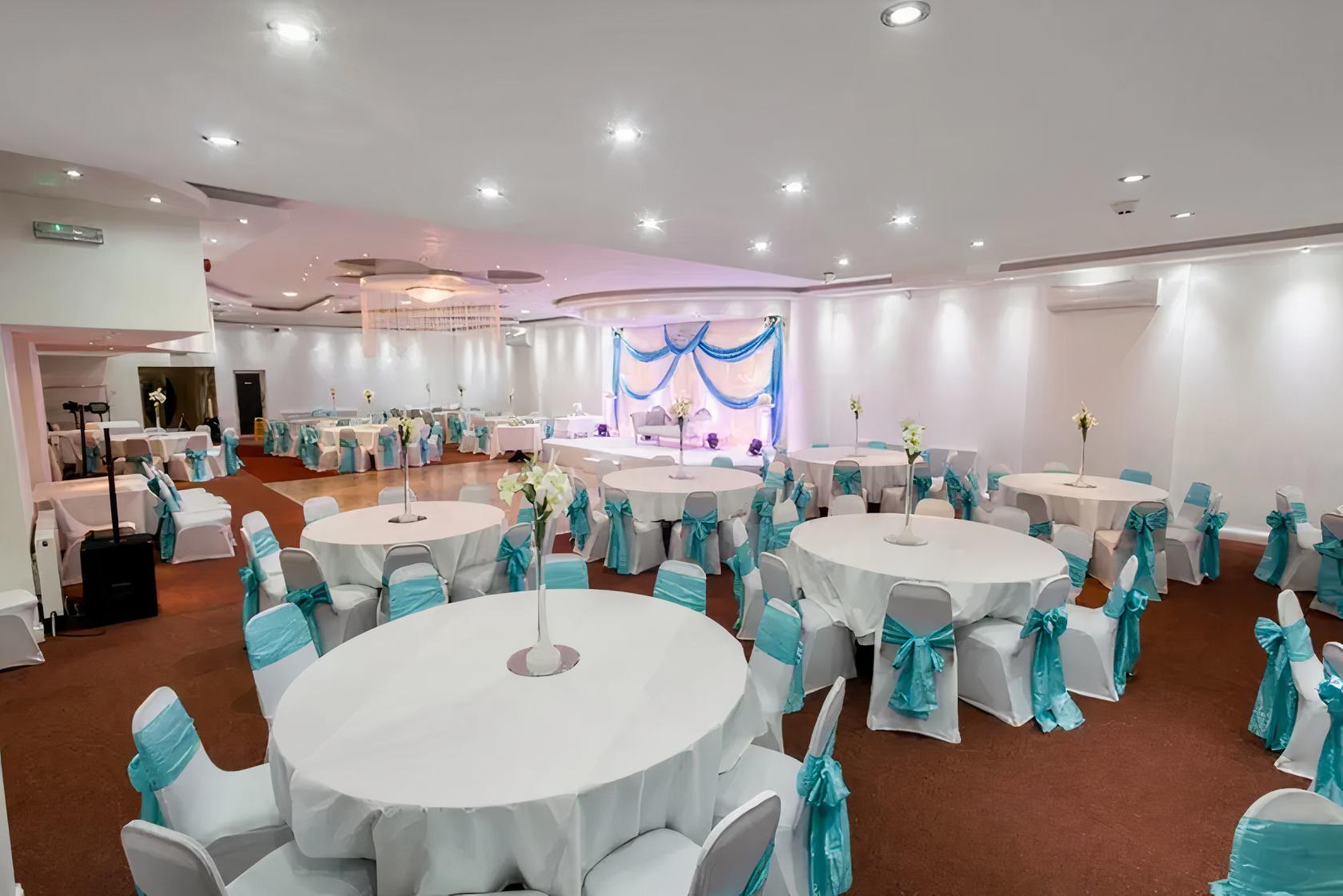 A banqueting hall with round tables covered in white linens and teal chair sashes at Elegance Q Banquet.