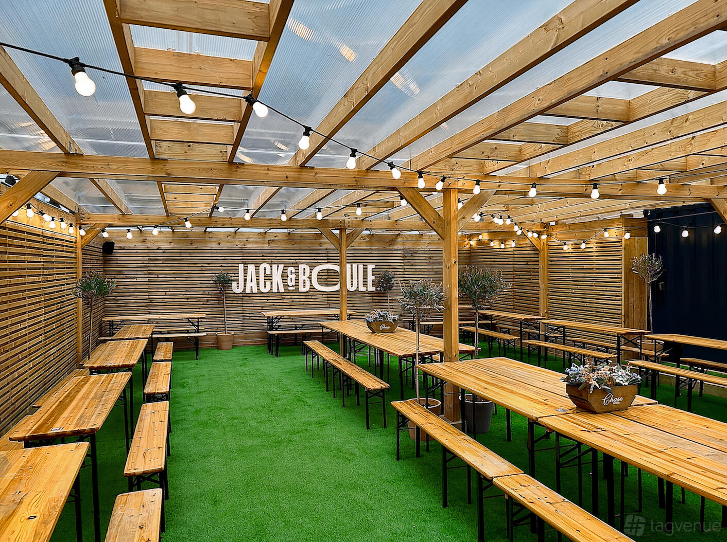 A bar with wooden picnic tables, string lights, and artificial grass under a clear roof at Boulebar South Bank.