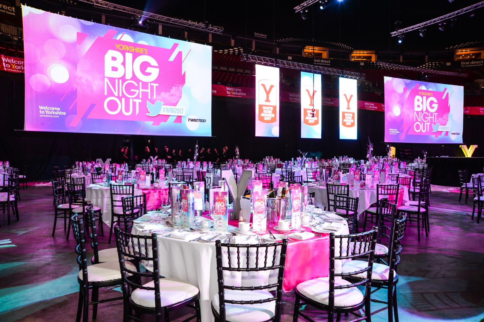 An auditorium with round banquet tables, pink décor accents, and large projection screens at first direct bank area - Leeds.