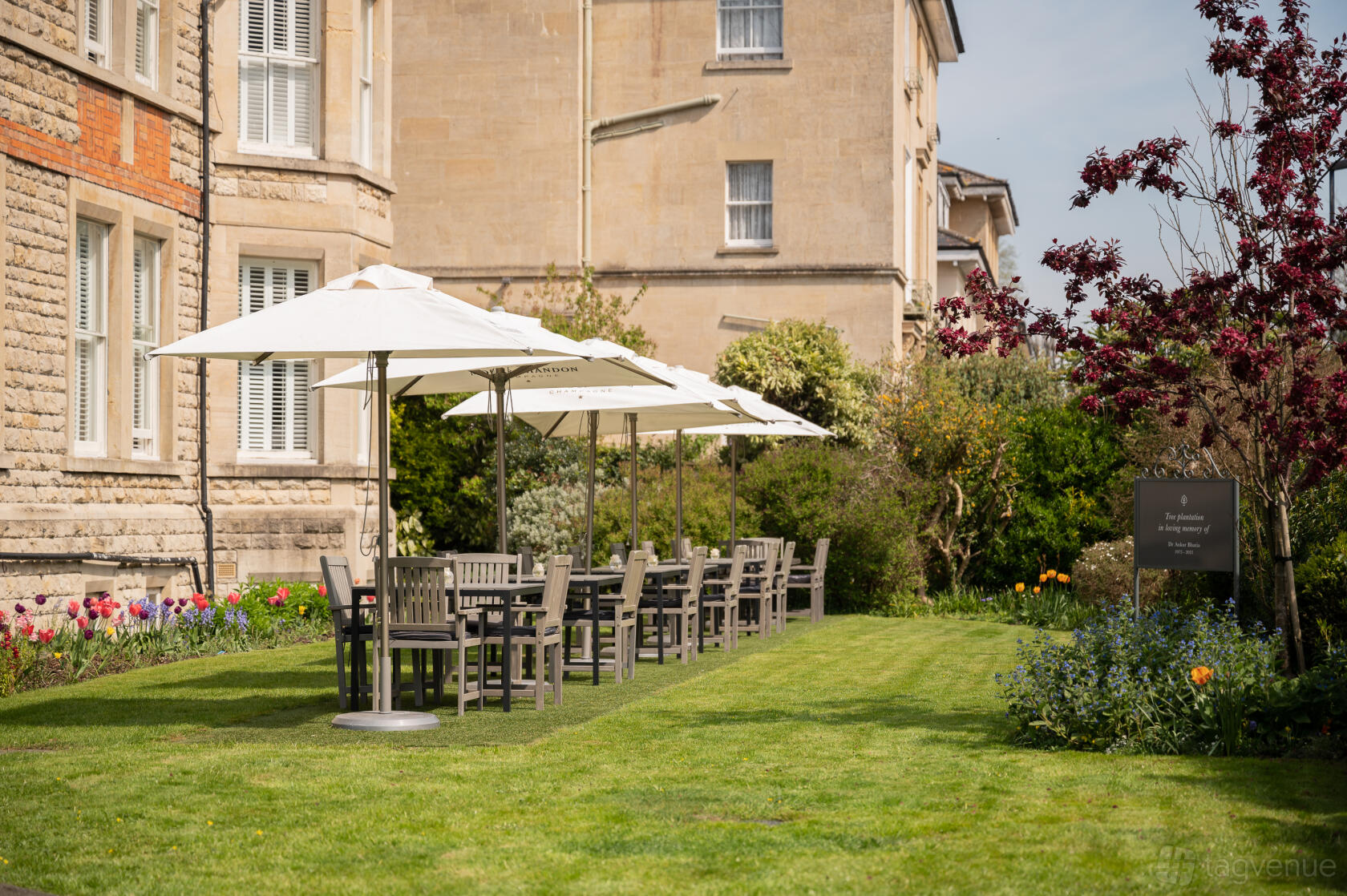 A hotel garden with wooden tables and chairs shaded by white umbrellas on a manicured lawn at The Roseate Villa Bath.