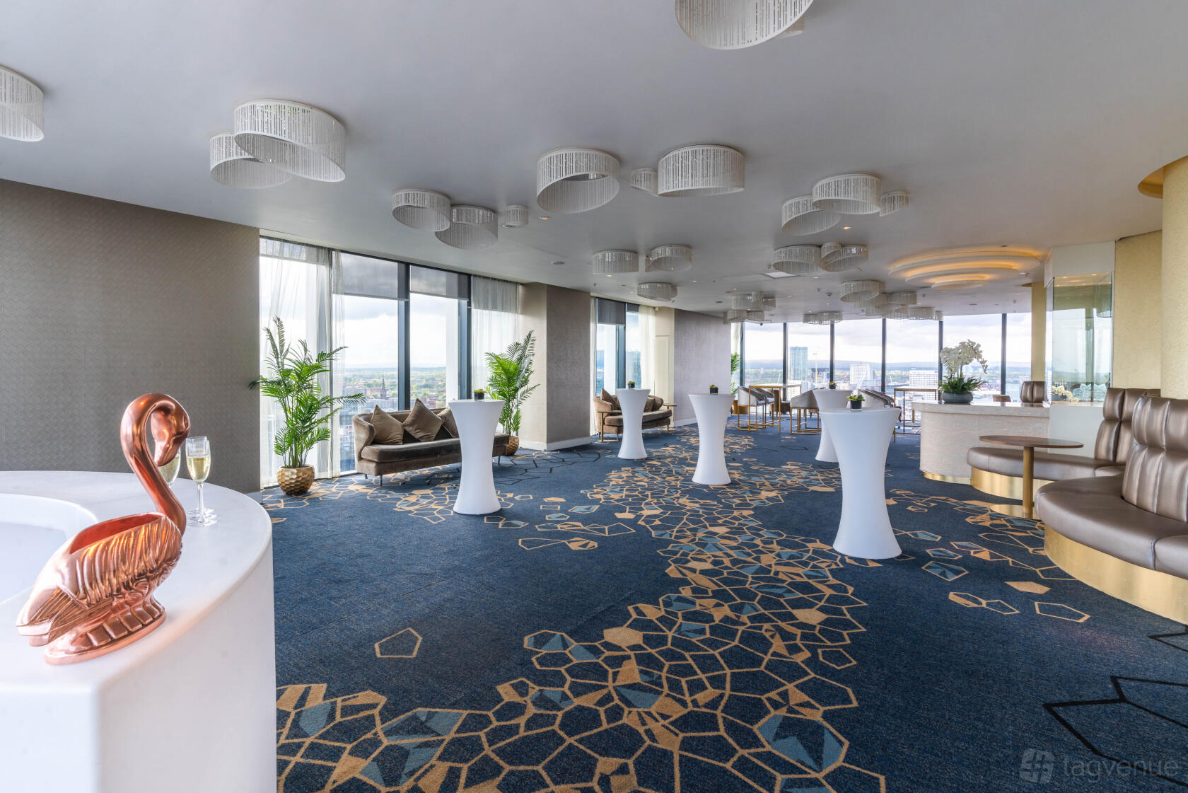 A cocktail bar with floor-to-ceiling windows, patterned carpeting, and white cocktail tables at Cloud 23.