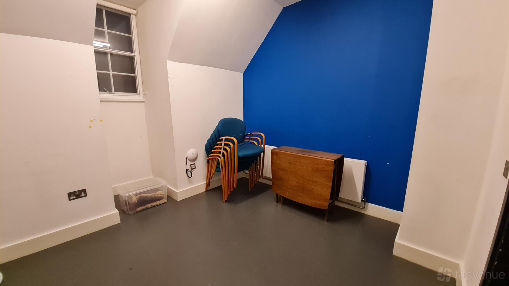 A theatre meeting room with a bold blue accent wall, stacked chairs, a folding table, and a small window at Bush Theatre.