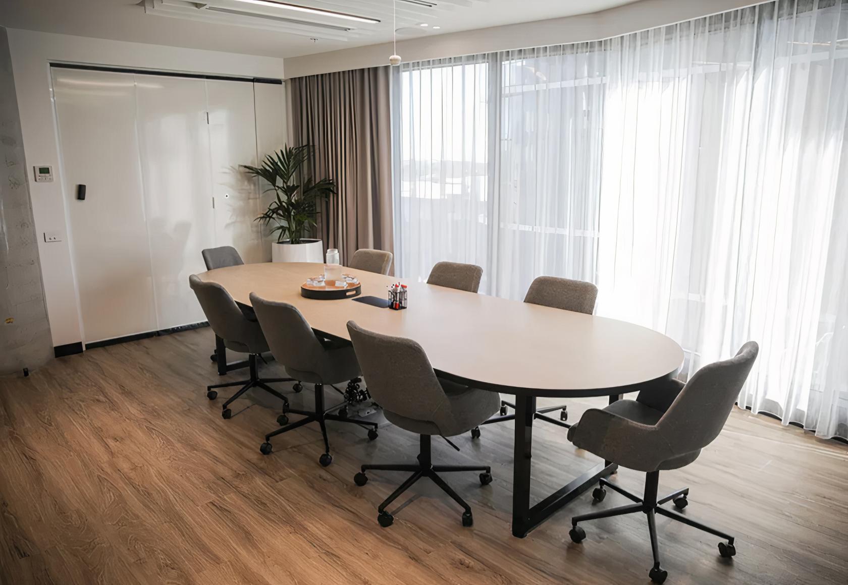 A meeting room with a long oval table, grey swivel chairs, and floor-to-ceiling sheer curtains at Hexa Space Coworking.