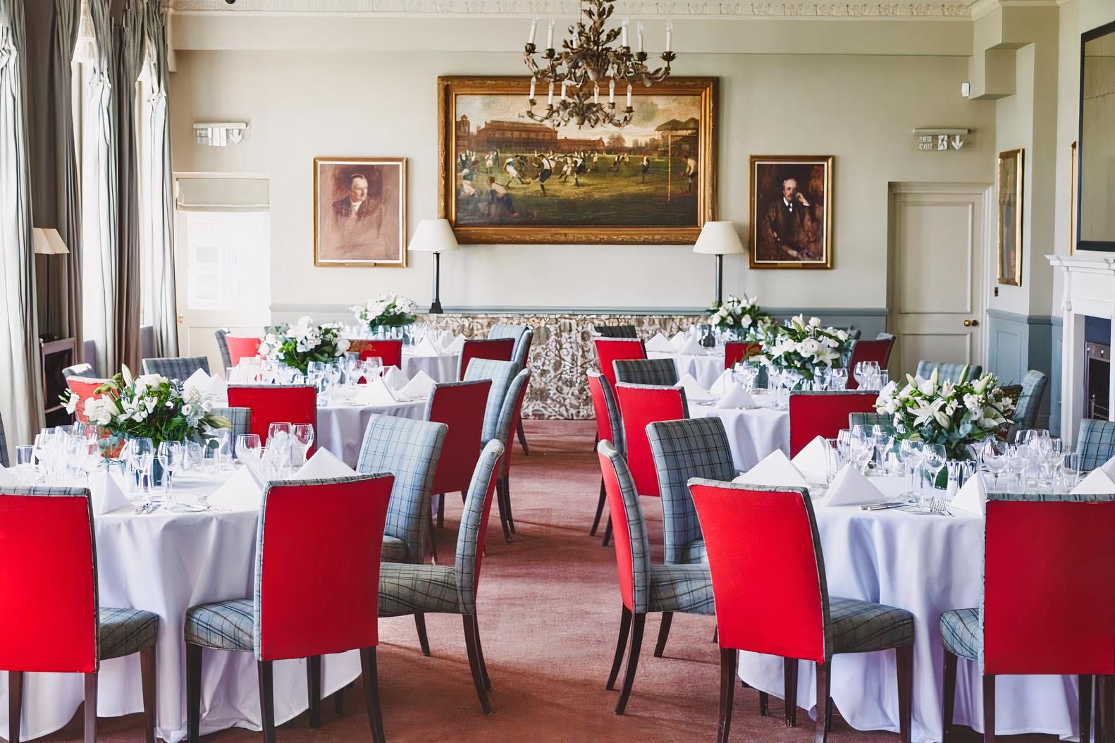 A function room with round tables covered in white linens, red chairs, and classic paintings at The Queen's Club.