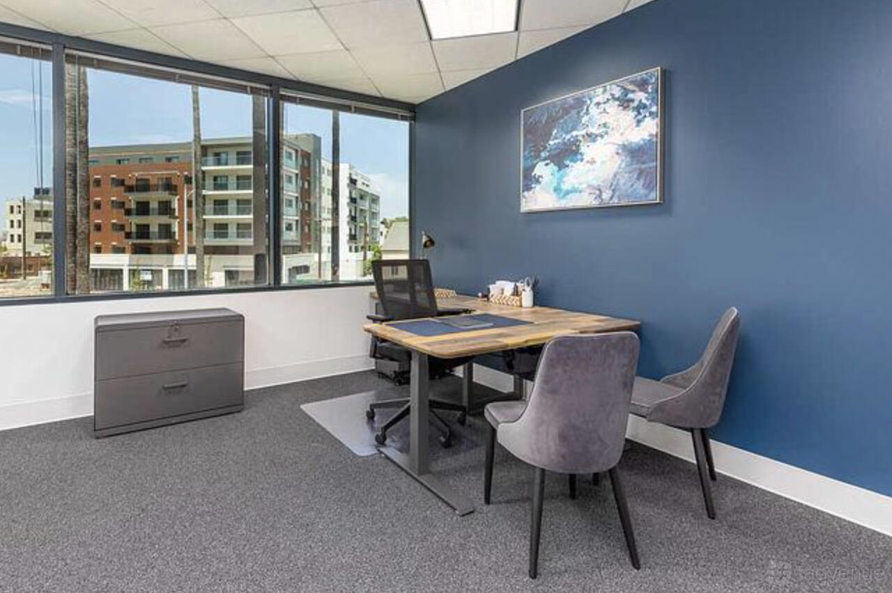A coworking space with a wooden desk, two gray chairs, blue accent wall, and large windows at Premier Workspaces - Executive Tower.
