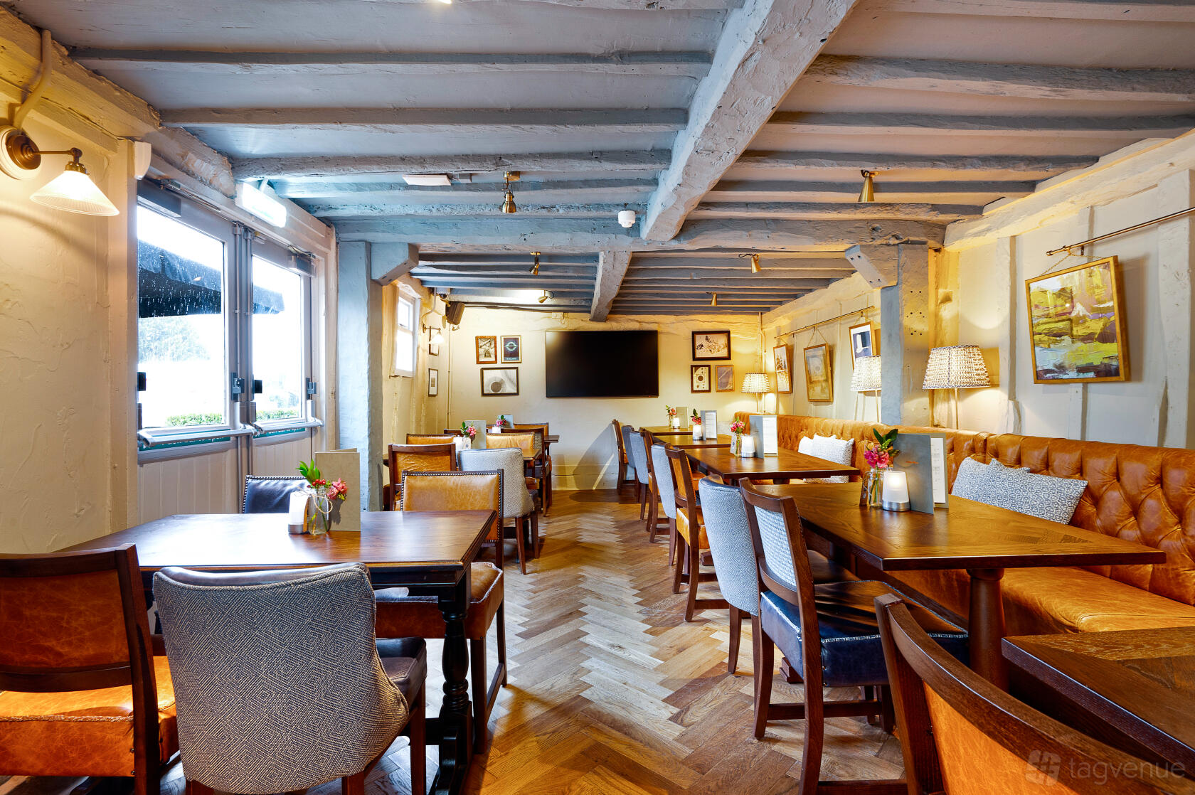 A pub with wooden tables, leather banquette seating, framed artwork, and herringbone floors at The King Harry.