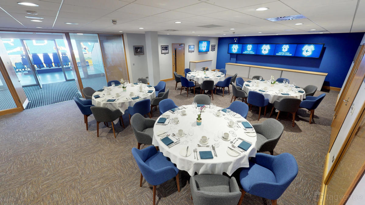 A stadium lounge with round tables set for dining, blue chairs, and wall-mounted screens at Cardiff City Stadium.