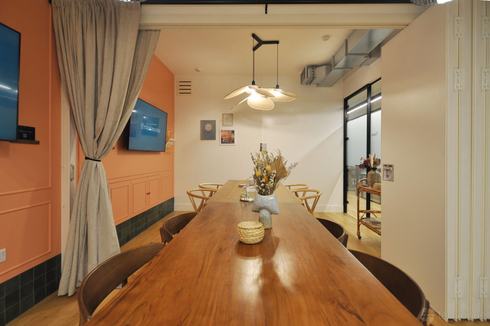 A meeting room with a long wooden table, modern chairs, wall-mounted screens, and pendant lighting at Missionworks.