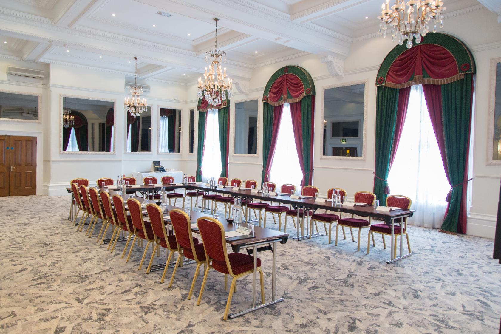 A banqueting hall with chandeliers, arched windows draped in red and green curtains, and U-shaped conference tables at Bristol Grand Hotel by Sunday.